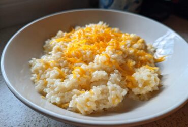 A close-up of a white bowl filled with creamy, amazing cheesy rice topped generously with melted shredded cheddar cheese.