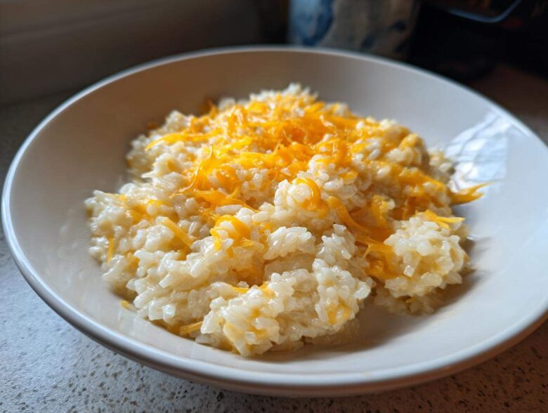 A close-up of a white bowl filled with creamy, amazing cheesy rice topped generously with melted shredded cheddar cheese.