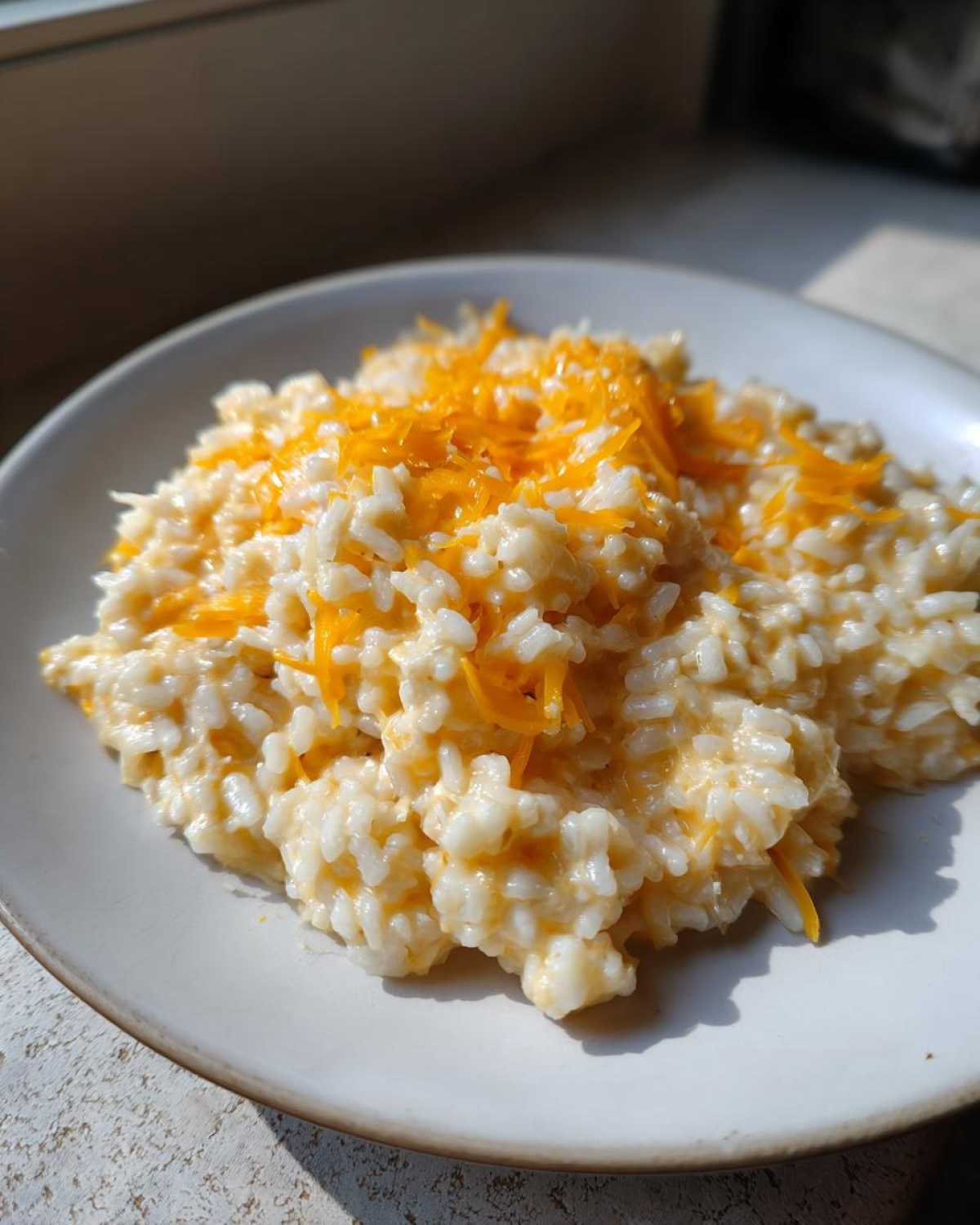 A serving of creamy cheesy rice topped with shredded orange cheddar cheese on a light plate.