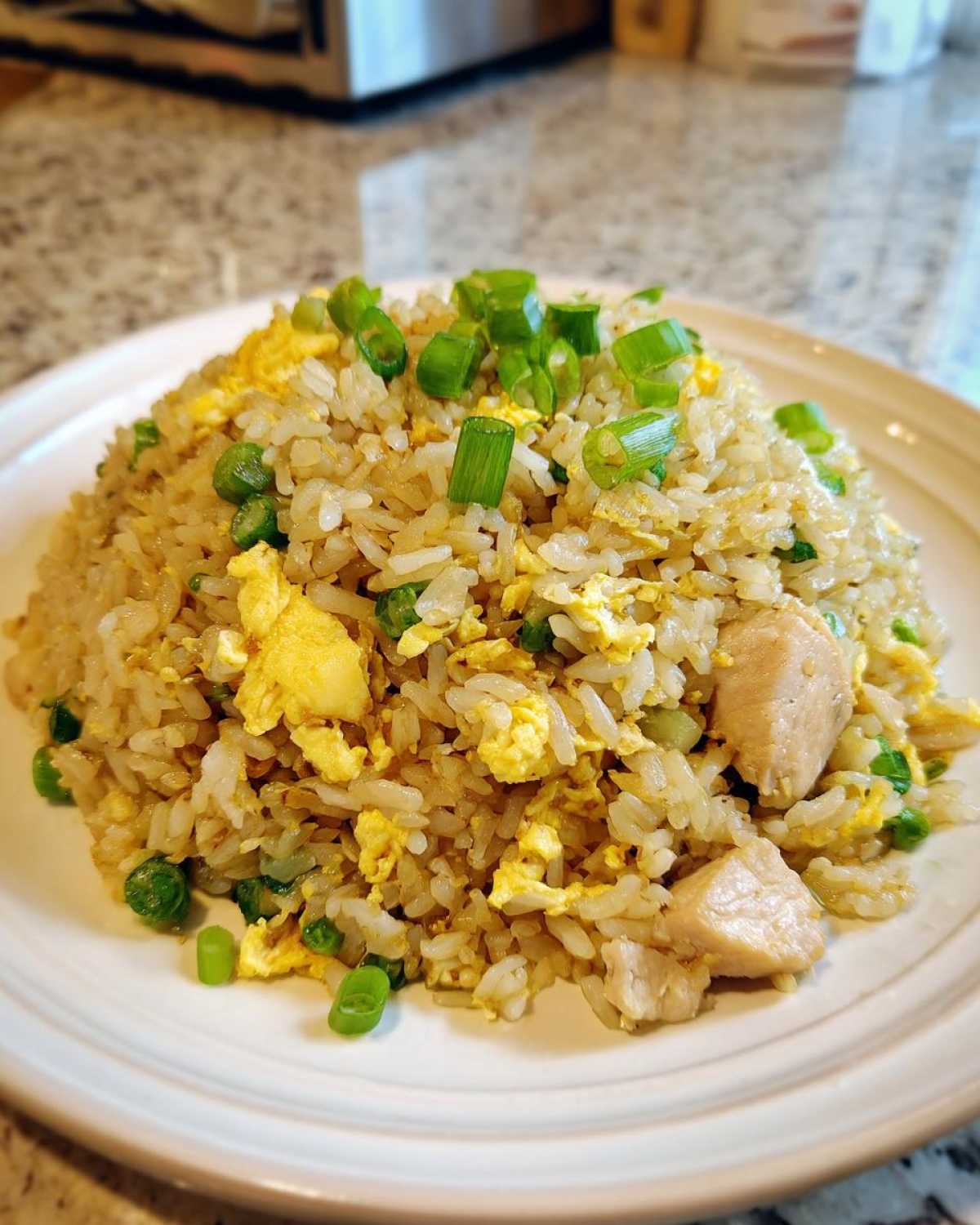 A mound of homemade chicken fried rice with scrambled egg, peas, and green onions on a white plate.