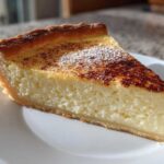 A thick slice of creamy egg custard tart dusted with cinnamon and powdered sugar on a white plate.