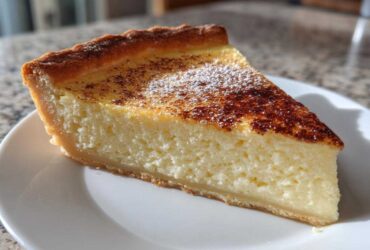 A thick slice of creamy egg custard tart dusted with cinnamon and powdered sugar on a white plate.