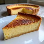 A close-up of a slice of creamy egg custard tart with a beautifully browned, slightly caramelized top, sitting on a white plate.