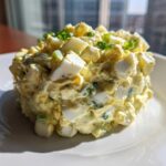 A molded mound of creamy, chunky egg salad recipe garnished with fresh chives on a white plate.