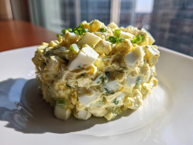 A molded mound of creamy, chunky egg salad recipe garnished with fresh chives on a white plate.