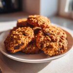 A close-up of several golden brown oatmeal cookie treats piled on a white plate, highlighted by natural sunlight.