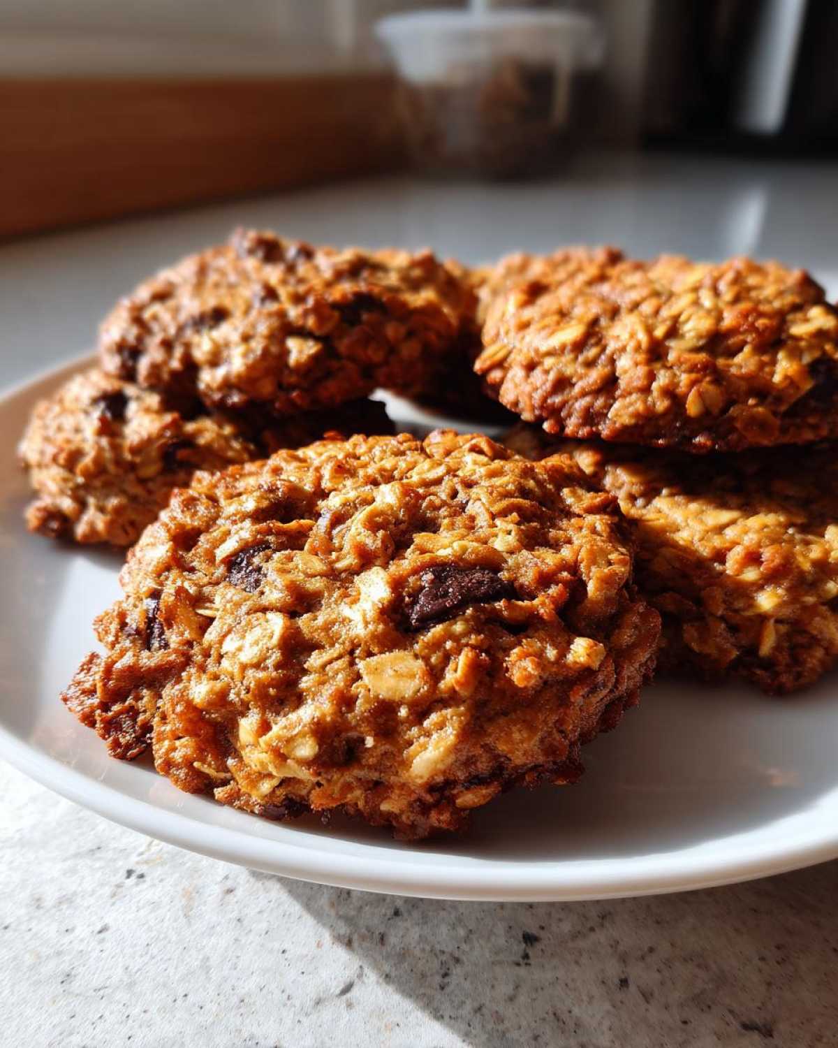 A close-up of several golden brown oatmeal cookie treats stacked on a white plate, catching warm sunlight.