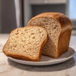 A freshly baked loaf of whole wheat bread with one thick slice leaning against it on a white plate.