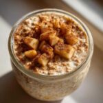 Close-up of apple cinnamon overnight oats in a glass jar, topped with diced apples and a generous dusting of cinnamon.