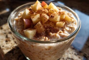 A close-up of creamy apple cinnamon overnight oats topped with diced apples and a dusting of cinnamon in a clear glass jar.