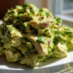 A mound of chunky avocado chicken salad, featuring visible pieces of chicken, mashed avocado, and red onion, served on a white plate.