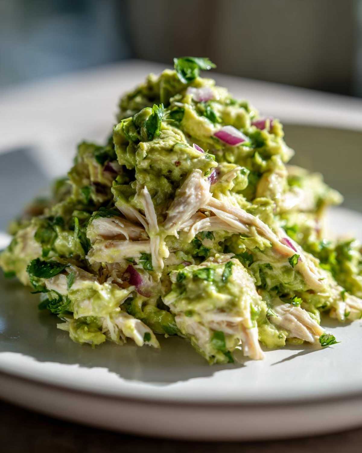 A mound of creamy avocado chicken salad mixed with red onion and cilantro served on a white plate.