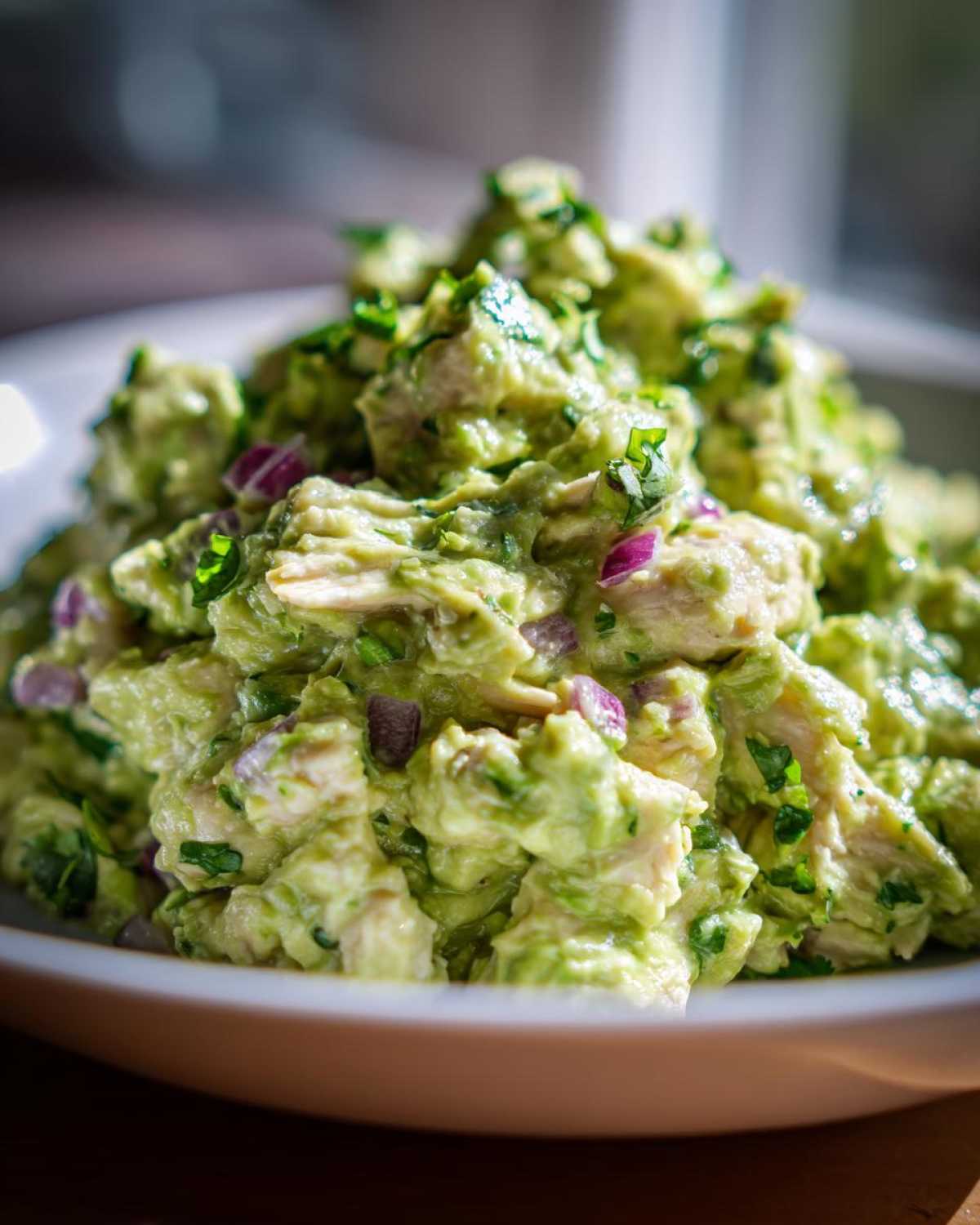A close-up, bright shot of creamy avocado chicken salad mixed with chunks of chicken and diced red onion, topped with cilantro.