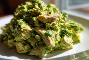 A mound of chunky avocado chicken salad, featuring visible pieces of chicken, mashed avocado, and red onion, served on a white plate.