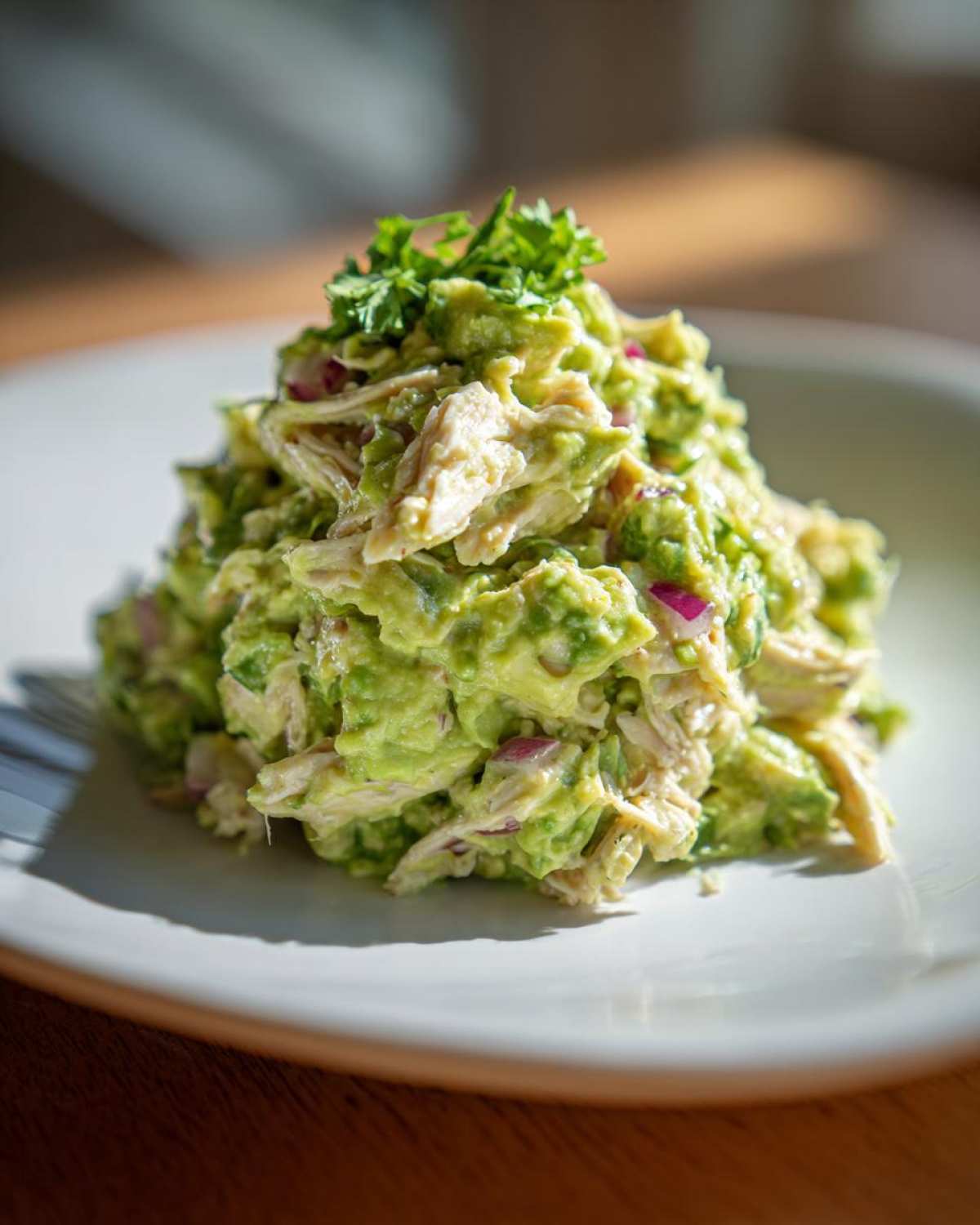 A mound of creamy avocado chicken salad mixed with red onion, topped with fresh parsley, served on a white plate.