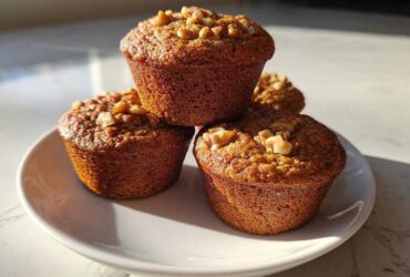 A stack of four golden brown banana nut muffins topped with walnuts, sitting on a white plate in bright sunlight.