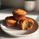 Three golden-brown banana protein muffins stacked on a white plate, one with a bite taken out showing the texture.