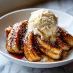 Close-up of caramelized bananas foster topped with a scoop of vanilla ice cream in a white bowl.
