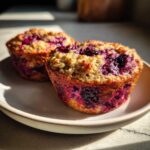 Two freshly baked berry protein muffins with deep purple fruit visible on a light ceramic plate.