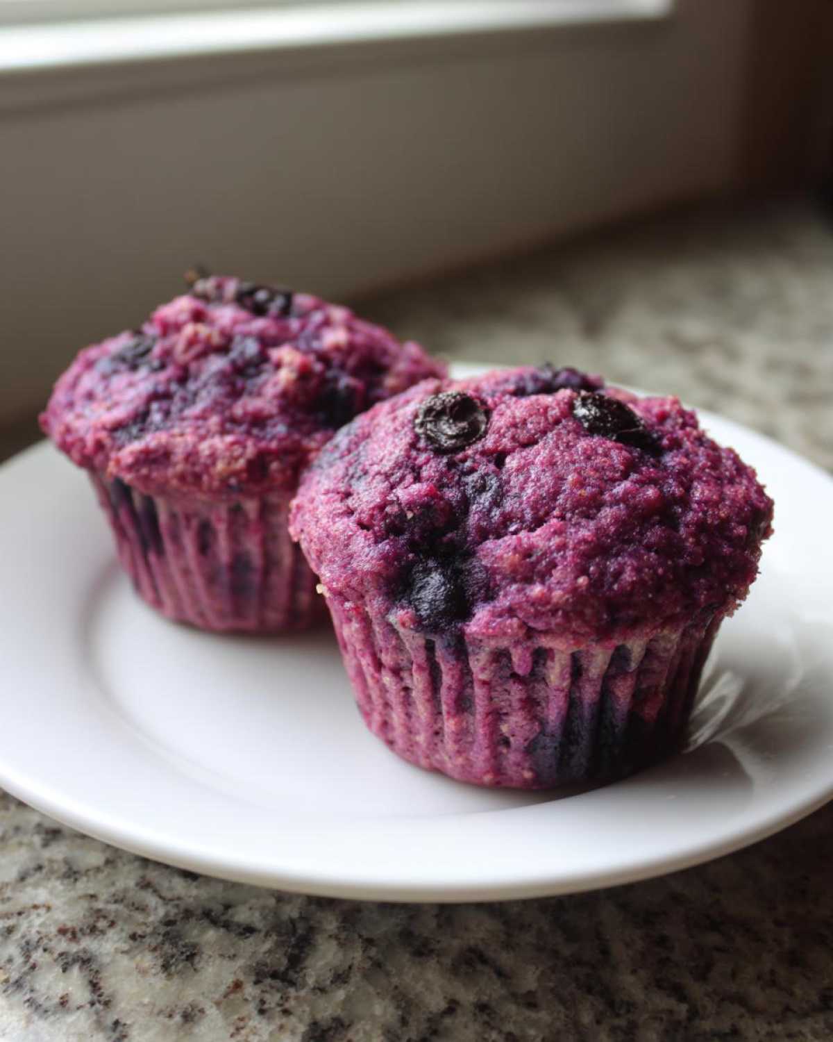 Two vibrant purple blueberry protein muffins displayed on a white plate near a window.