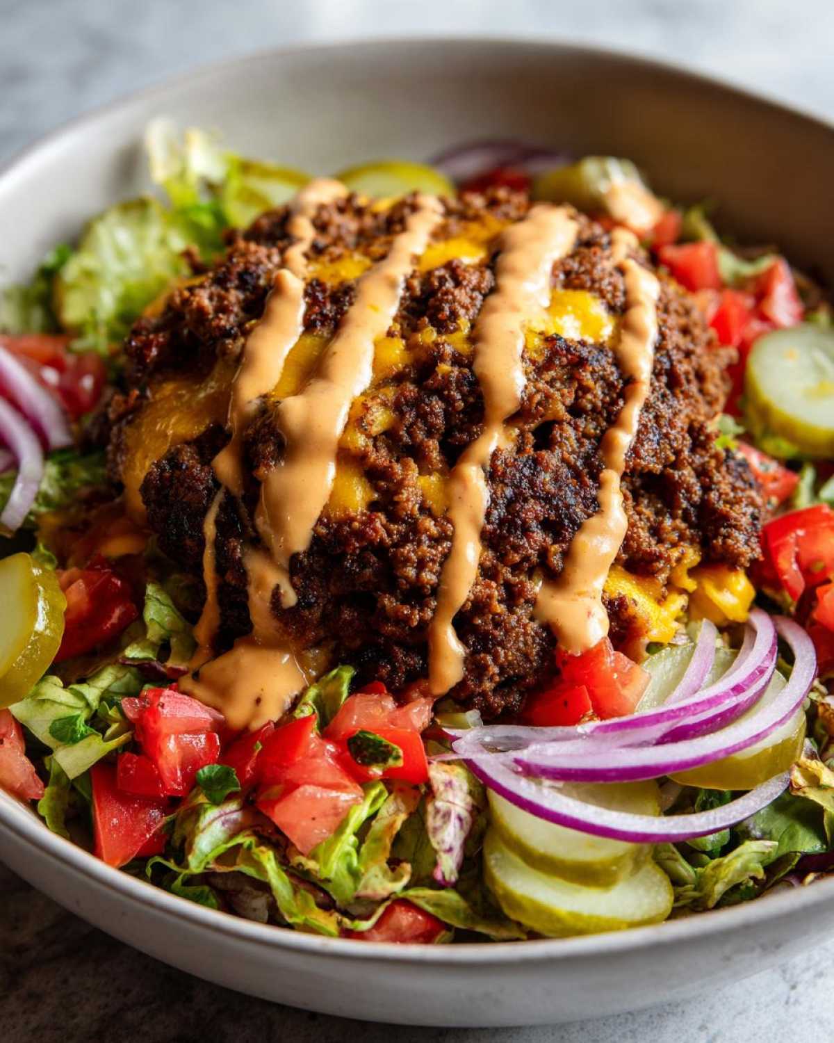 A close-up of a cheeseburger bowl featuring seasoned ground beef, melted cheese, and a drizzle of sauce over lettuce, tomatoes, pickles, and red onion.