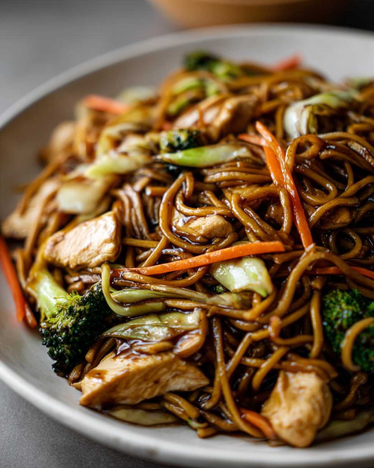 Close-up of a bowl filled with saucy chicken chow mein recipe featuring noodles, chicken pieces, broccoli, and carrots.