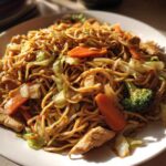 A close-up shot of a plate filled with homemade chicken chow mein recipe, featuring noodles, chicken pieces, carrots, and broccoli.