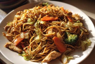 A close-up shot of a plate filled with homemade chicken chow mein recipe, featuring noodles, chicken pieces, carrots, and broccoli.