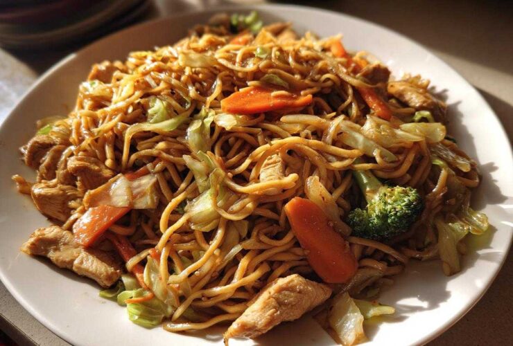 A close-up shot of a plate filled with homemade chicken chow mein recipe, featuring noodles, chicken pieces, carrots, and broccoli.
