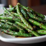 A close-up of glossy, stir-fried chinese green beans with garlic sauce and sesame seeds on a white plate.