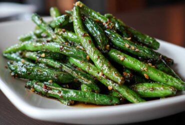 A close-up of glossy, stir-fried chinese green beans with garlic sauce and sesame seeds on a white plate.