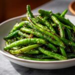 A close-up shot of vibrant, glossy chinese green beans with garlic sauce piled high in a white bowl.