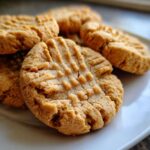 A close-up shot of several golden brown peanut butter cookies stacked on a white plate, showing the classic crisscross fork marks.