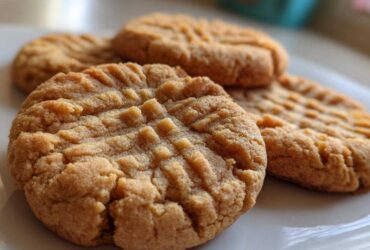 Close-up of several golden brown peanut butter cookies featuring the classic criss-cross fork pattern on a white plate.