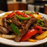 Close-up of sizzling steak fajitas featuring strips of seasoned beef mixed with vibrant red, green, and yellow bell peppers and onions.