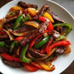 Close-up of sizzling steak fajitas featuring strips of seasoned beef mixed with vibrant red, green, and yellow bell peppers and onions.