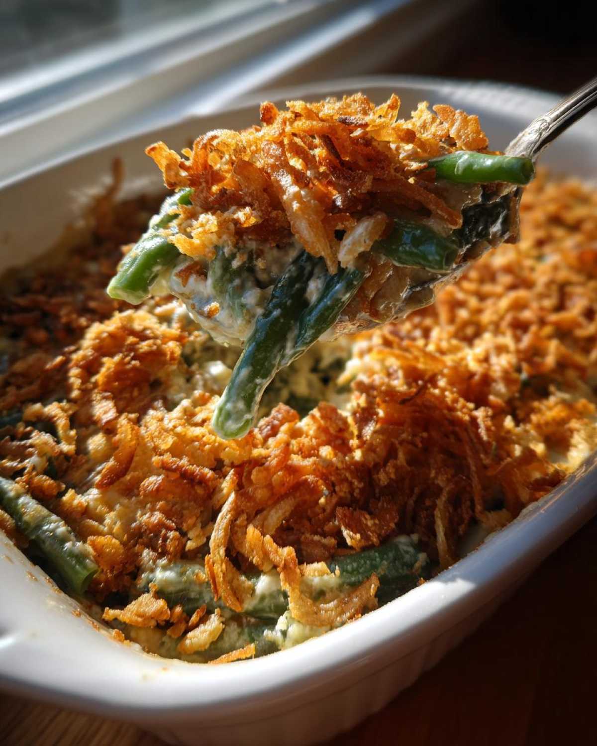 A spoonful of creamy green bean casserole topped with crispy fried onions being lifted from a white baking dish.