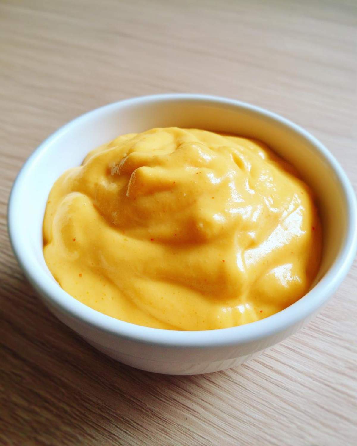Close-up of rich, smooth, orange nacho cheese sauce served in a small white bowl on a wooden surface.