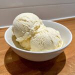 Three scoops of creamy, vanilla-colored protein ice cream served in a white bowl on a wooden surface.
