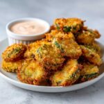 A pile of golden brown, crispy fried pickles served on a white plate with a small ramekin of dipping sauce.