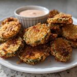 A plate piled high with golden brown, crispy fried pickles served with a small ramekin of creamy dipping sauce.
