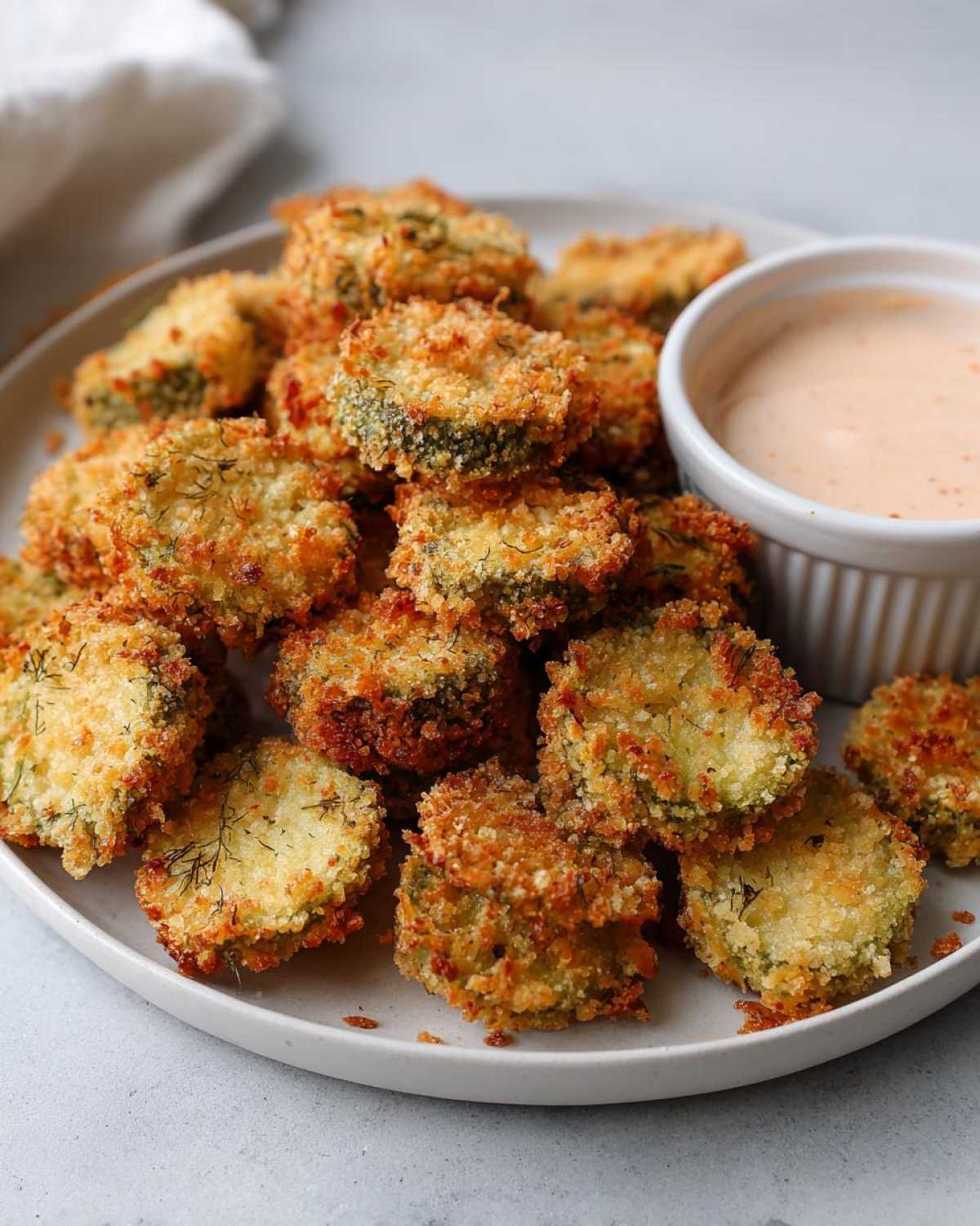 A plate piled high with golden brown, crispy Fried Pickles served next to a small ramekin of creamy dipping sauce.