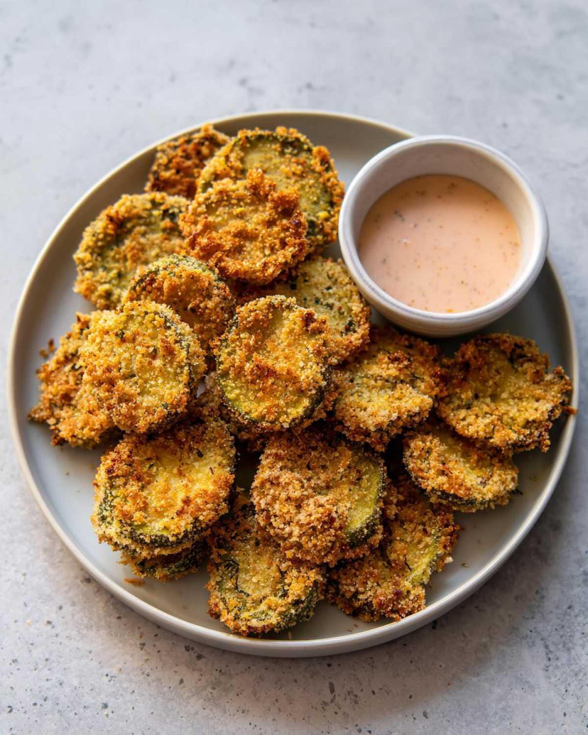 A plate piled high with golden brown, crispy Fried Pickles slices next to a small bowl of creamy dipping sauce.