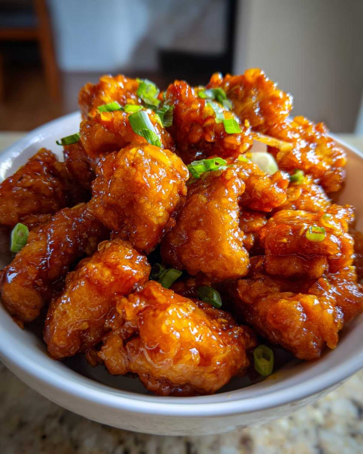 Close-up of crispy, glazed pieces of orange chicken recipe topped with sliced green onions in a white bowl.
