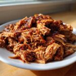 A generous serving of saucy, shredded crock pot pulled pork piled high on a white plate.