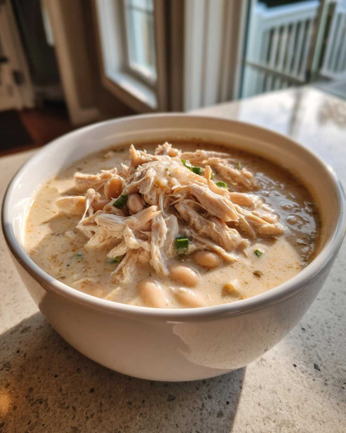 A close-up of a white bowl filled with creamy crockpot white chicken chili, topped with shredded chicken and green onions.