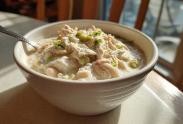 A close-up of a white bowl filled with creamy crockpot white chicken chili, topped with shredded chicken and green onions.