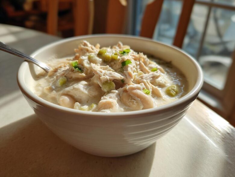 A close-up of a white bowl filled with creamy crockpot white chicken chili, topped with shredded chicken and green onions.