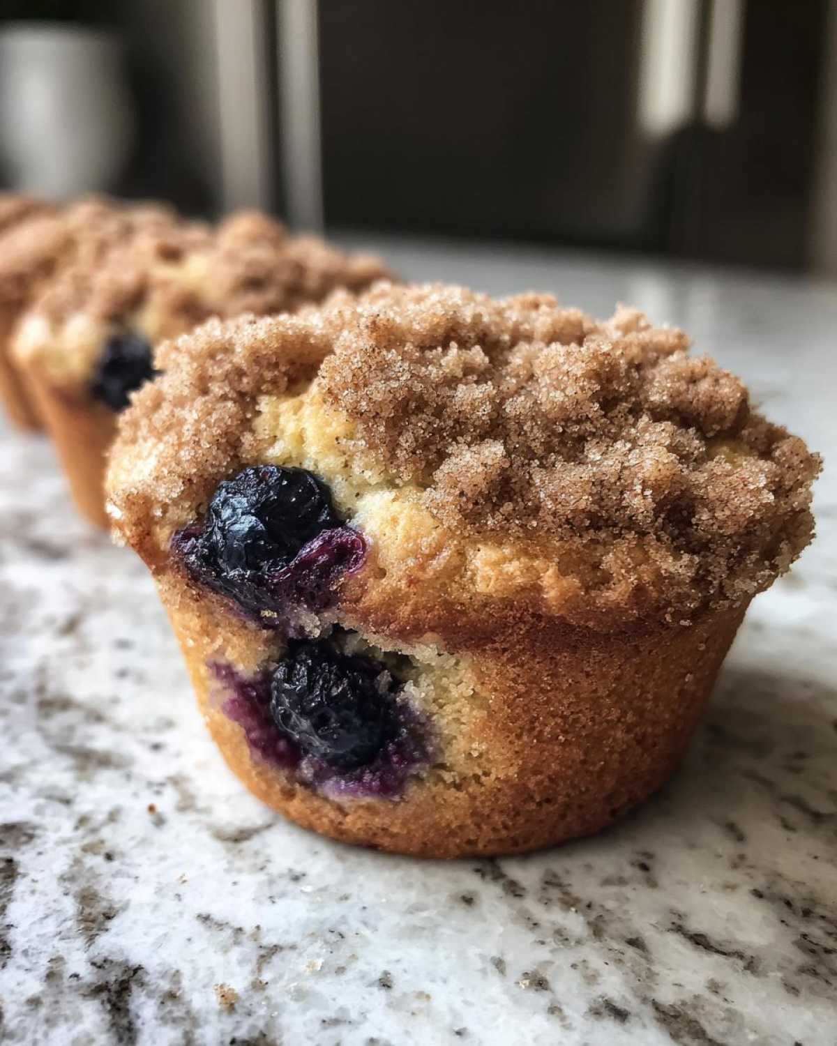 A close-up of a delicious blueberry muffin topped with cinnamon sugar crumble, sitting on a granite counter.