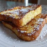 Two slices of golden brown easy french toast stacked and dusted with powdered sugar on a white plate.